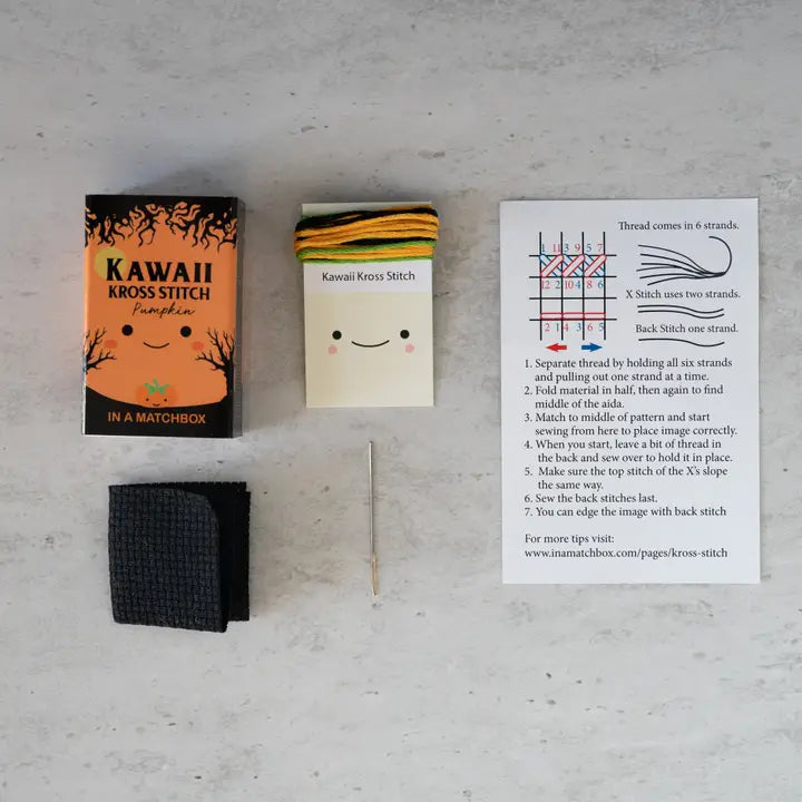 Kawaii Kross Stitch kit with a matchbox, fabric swatch, and instruction sheet on a gray surface.
