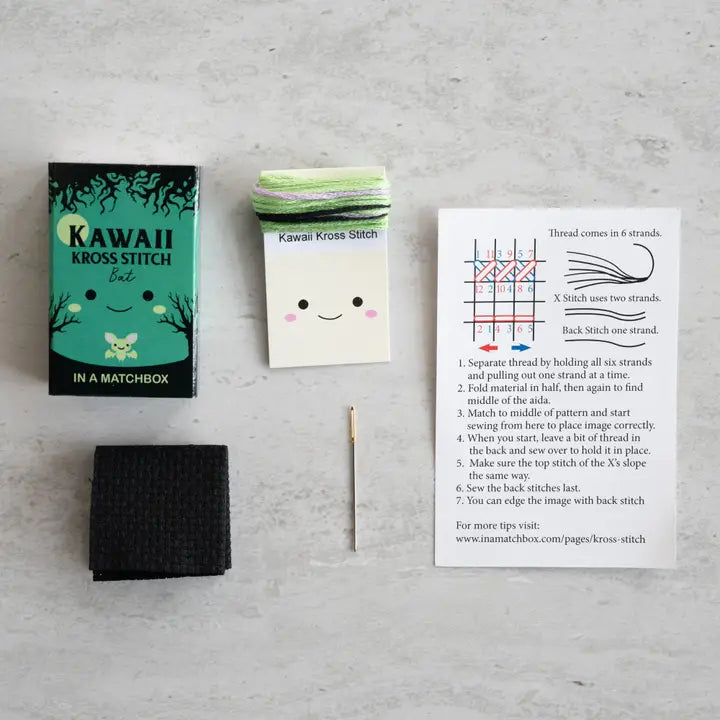 Kawaii Kross Stitch kit with a matchbox, black fabric, and stitch guide on a gray surface.