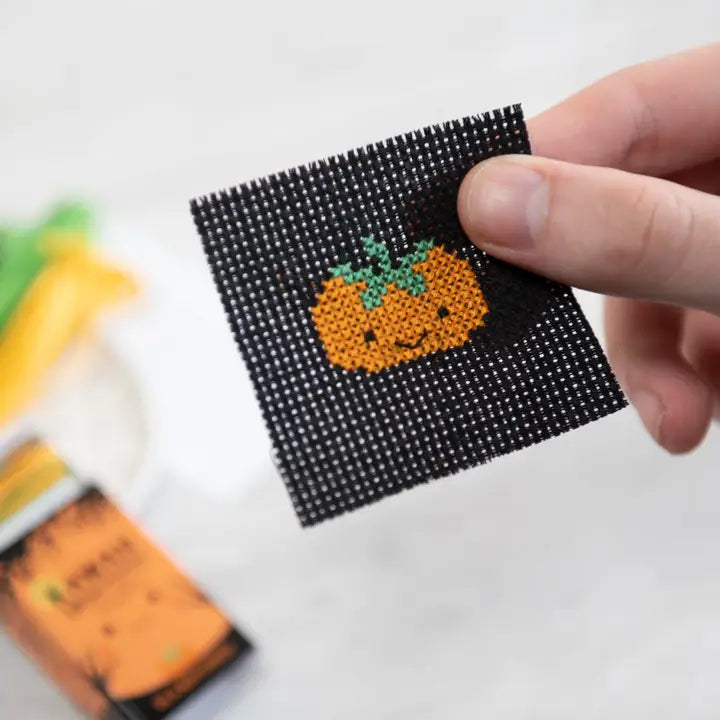 Hand holding a cross-stitched pumpkin design on a black fabric square with blurred craft supplies in the background.
