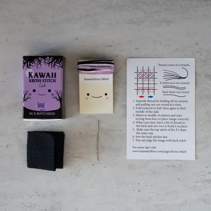Kawaii Kross Stitch kit with matchbox, fabric swatch, and instruction sheet on a gray surface