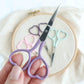 Hand holding a pair of purple scissors with embroidery hoop and heart-shaped scissors in the background