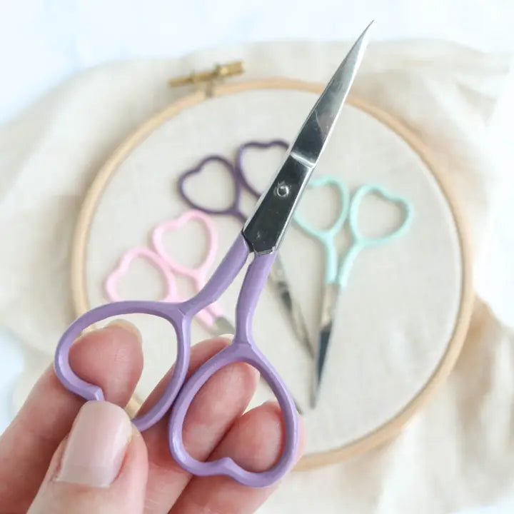 Hand holding a pair of purple scissors with embroidery hoop and heart-shaped scissors in the background
