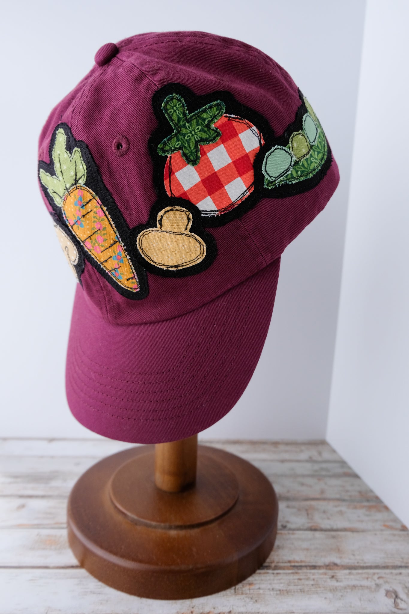 Maroon cap with vegetable patches on a wooden stand against a white wall.
