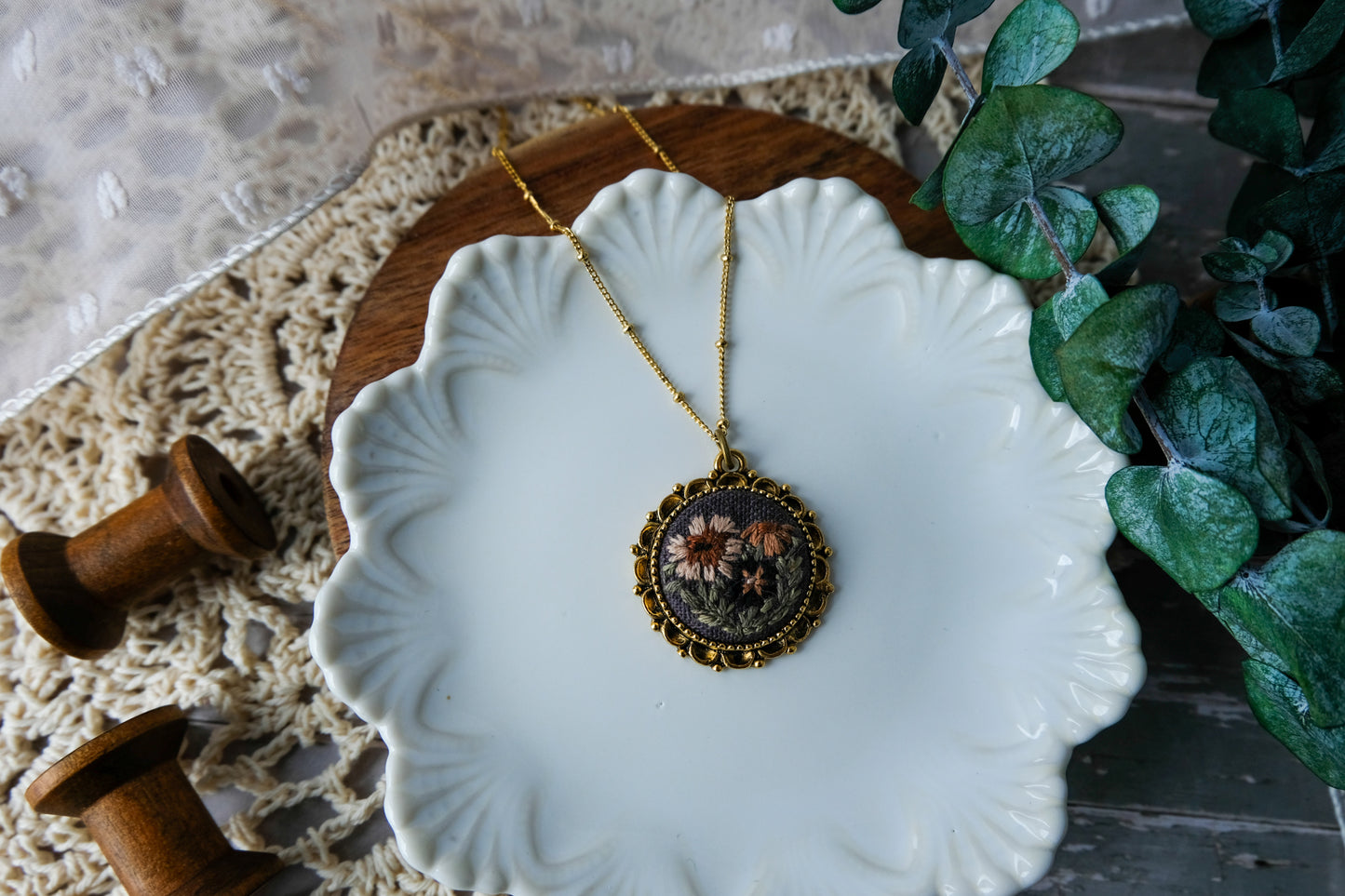 Necklace with a decorative pendant on a white dish, surrounded by greenery and wooden spools.
