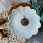Necklace with a decorative pendant on a white ceramic dish, surrounded by greenery and lace.