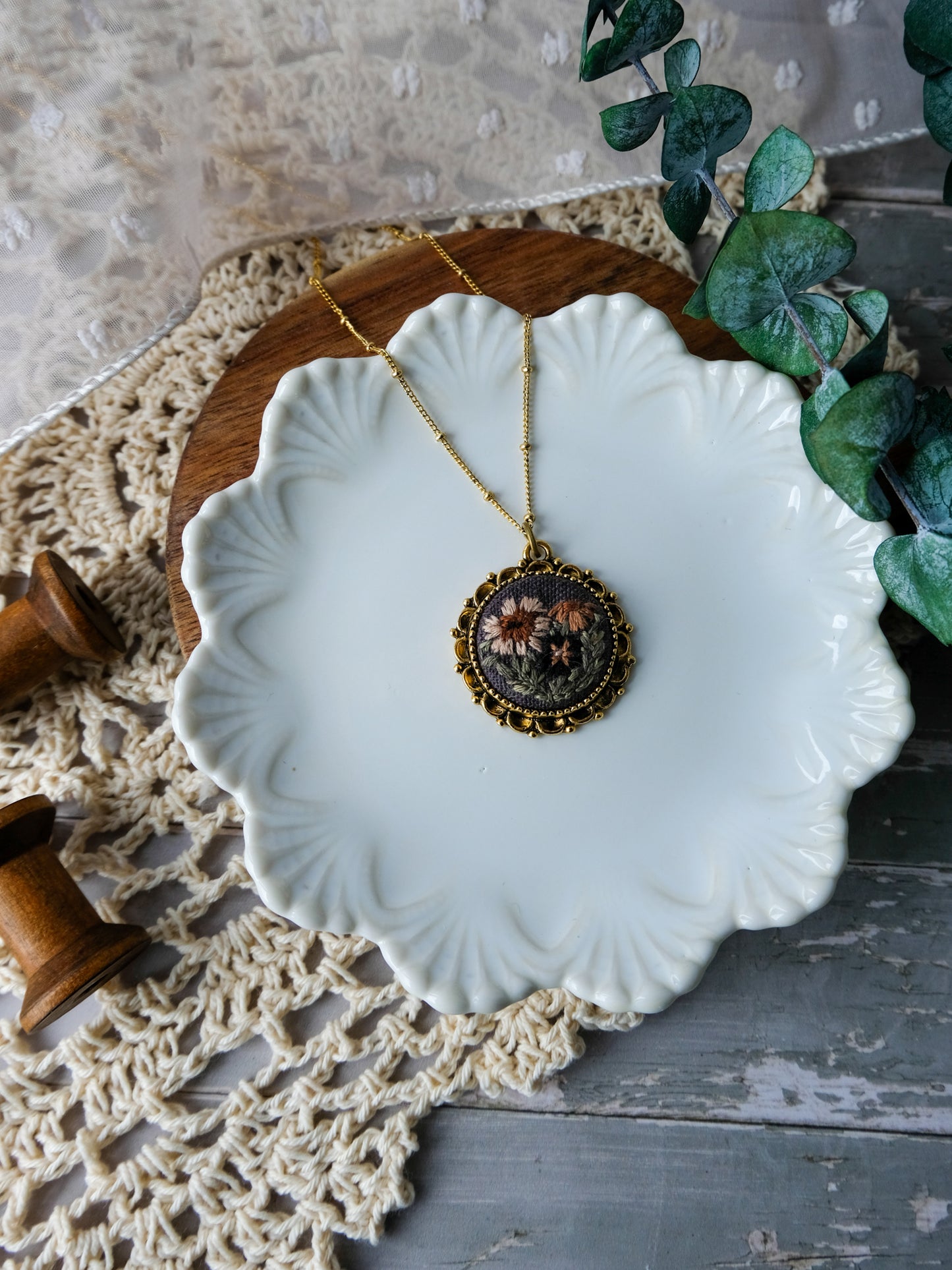 Necklace with a decorative pendant on a white ceramic dish, surrounded by greenery and lace.