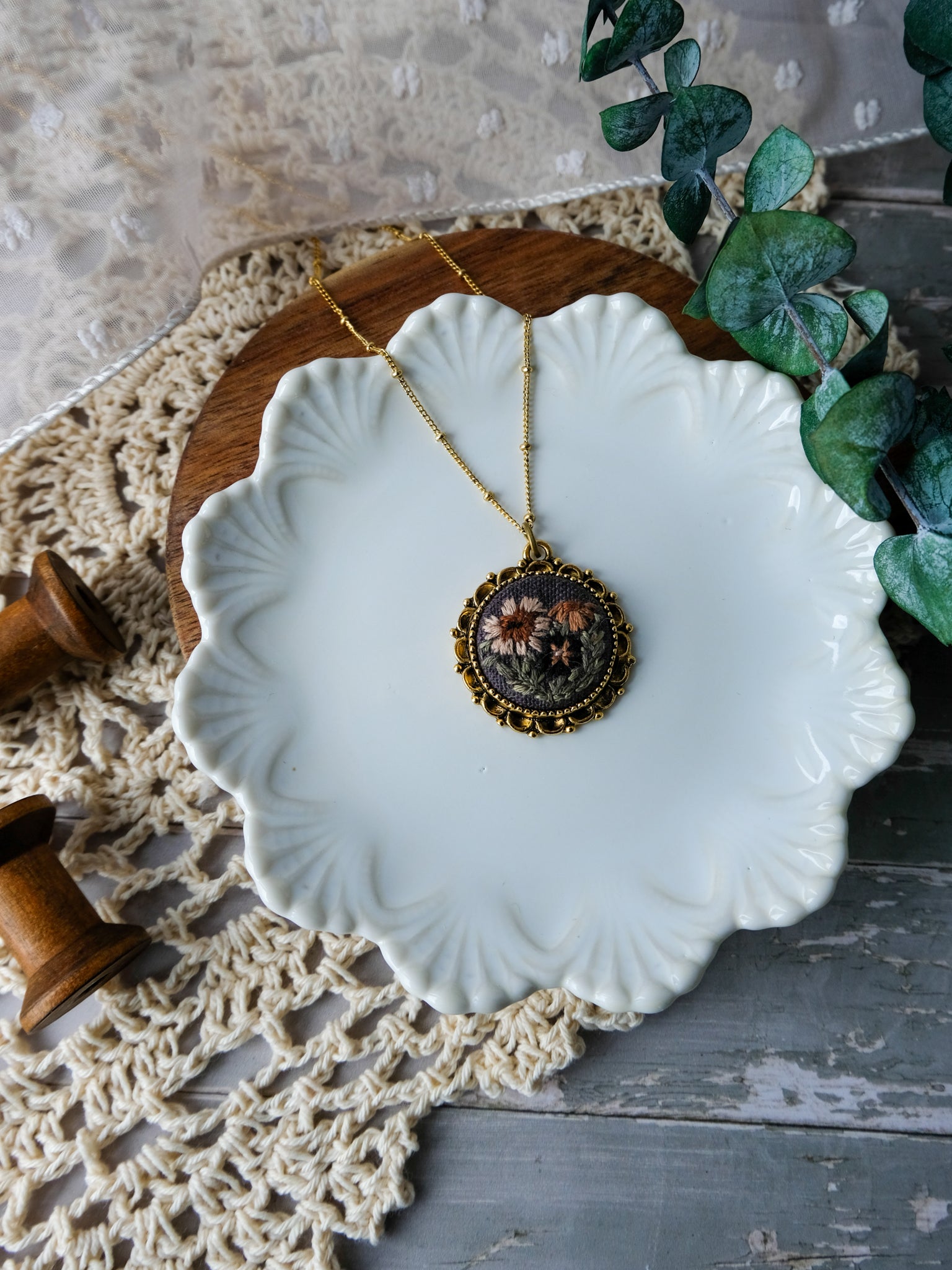 Necklace with a decorative pendant on a white ceramic dish, surrounded by greenery and lace.
