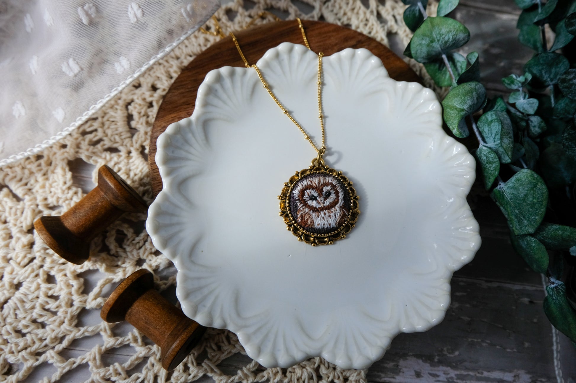 Jewelry dish with an owl pendant necklace on a textured surface with greenery.