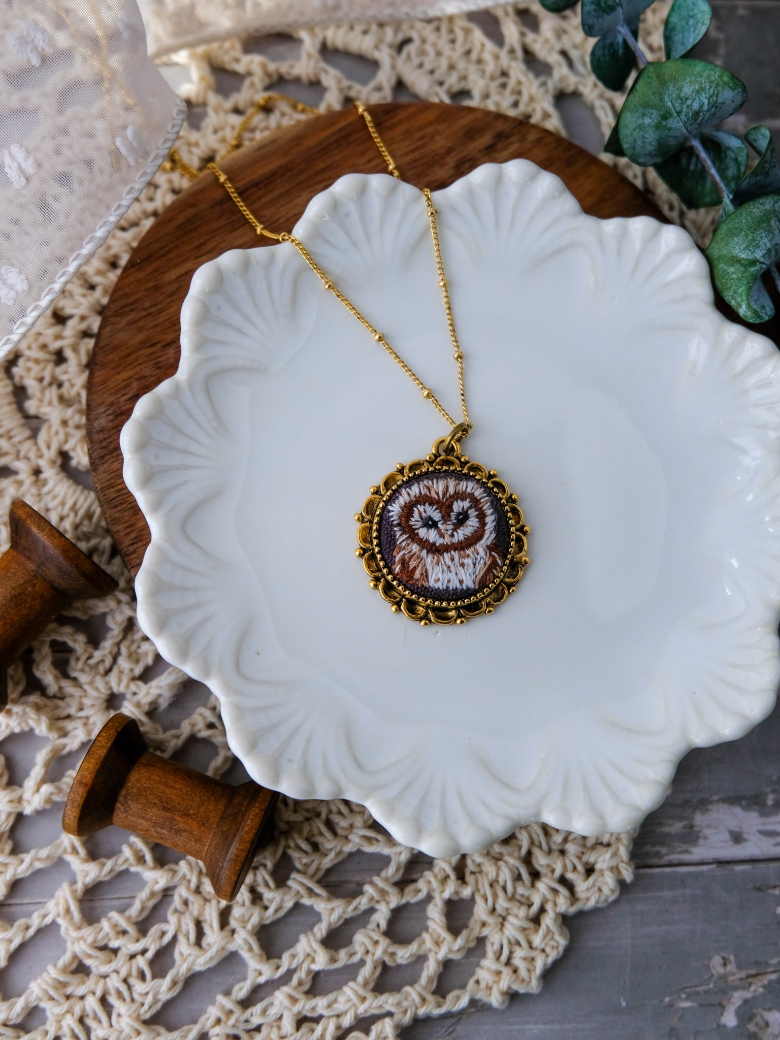 Necklace with an embroidered pendant on a white dish, wooden stand, and lace background