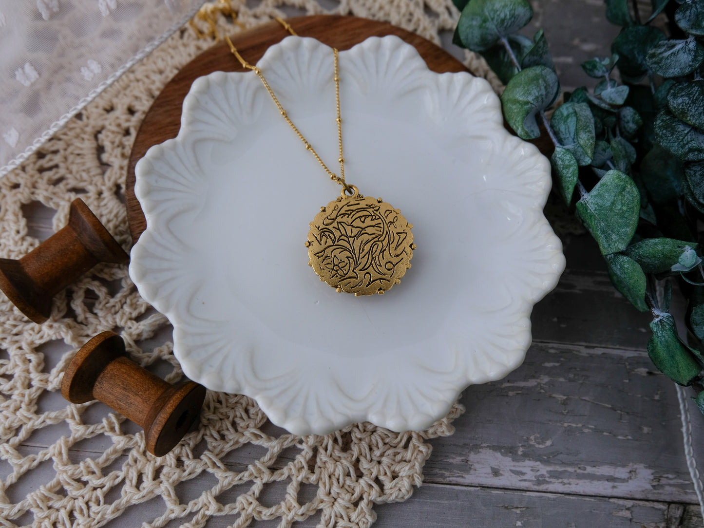 Gold necklace on a white ceramic dish with wooden spools and greenery in the background