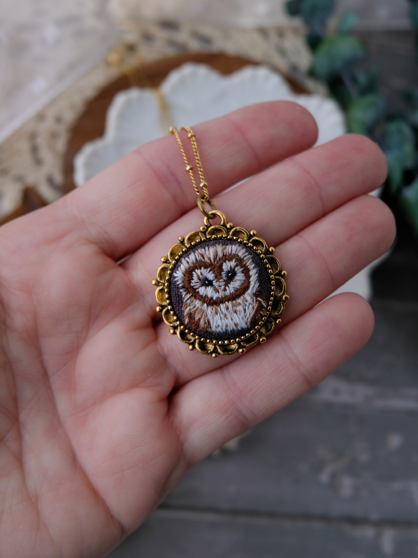 Hand holding a gold-framed embroidered pendant with an owl design.