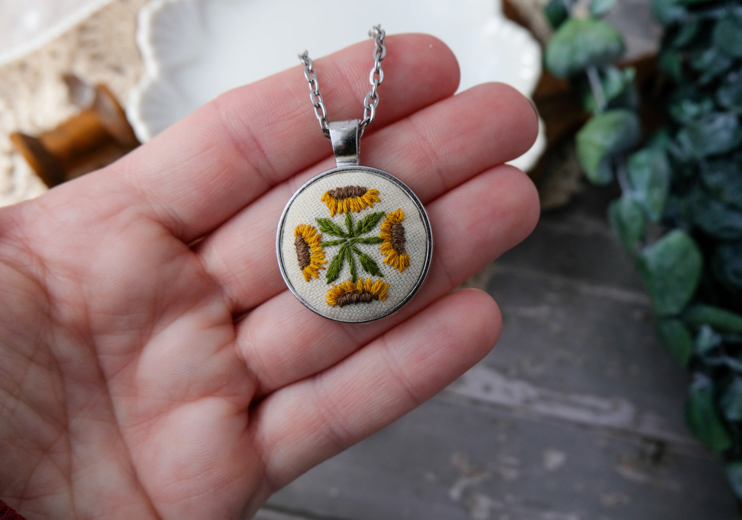 Hand holding a necklace with an embroidered pendant featuring a floral design.