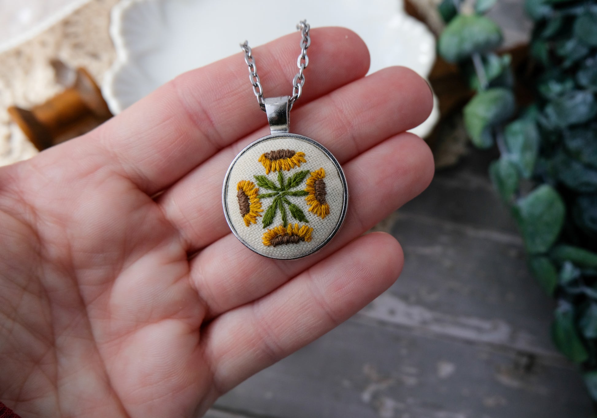 Hand holding a necklace with an embroidered pendant featuring a floral design.
