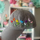 Gray cap with colorful embroidered flowers held in front of a blurred floral background