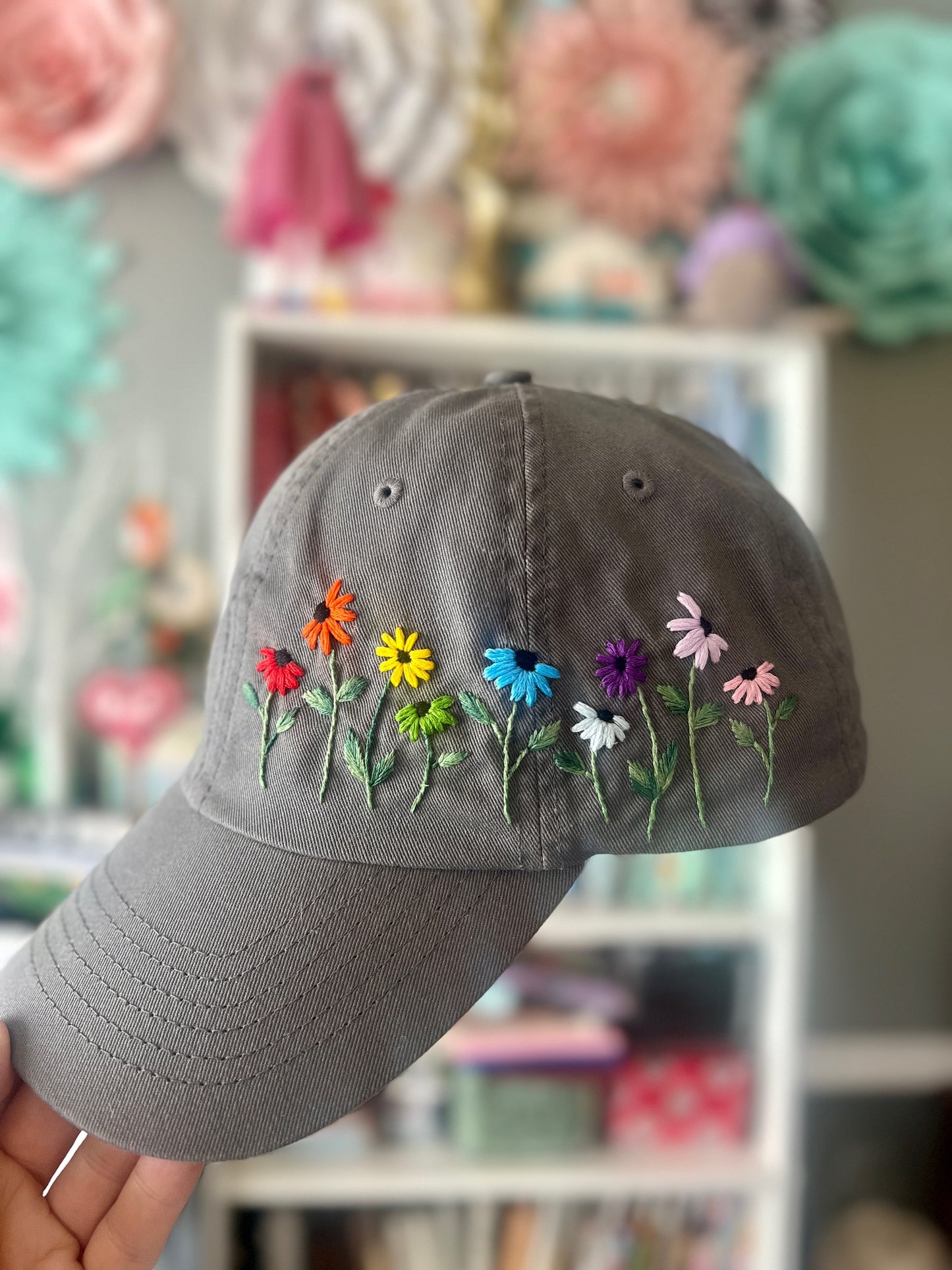 Gray cap with colorful embroidered flowers held in front of a blurred floral background