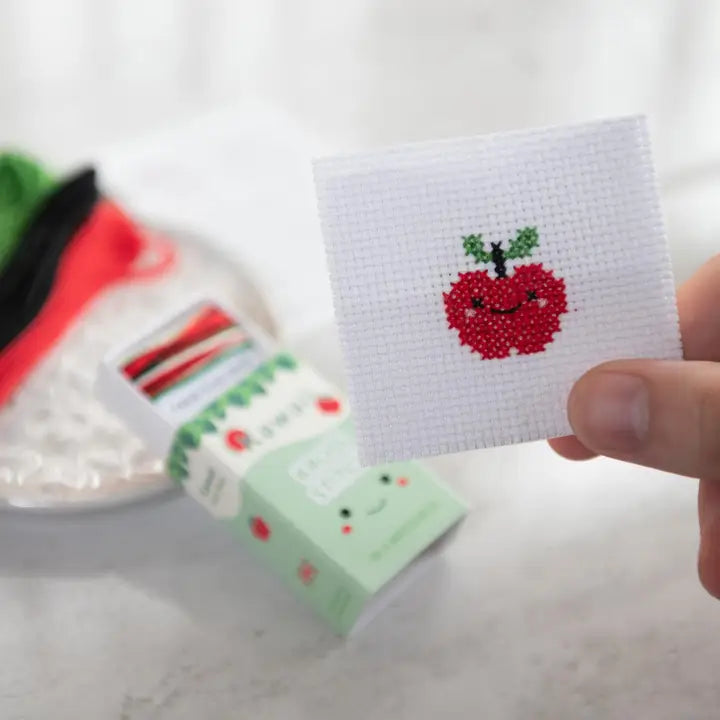 Hand holding a cross-stitched patch of a red apple with a smile on a white background.