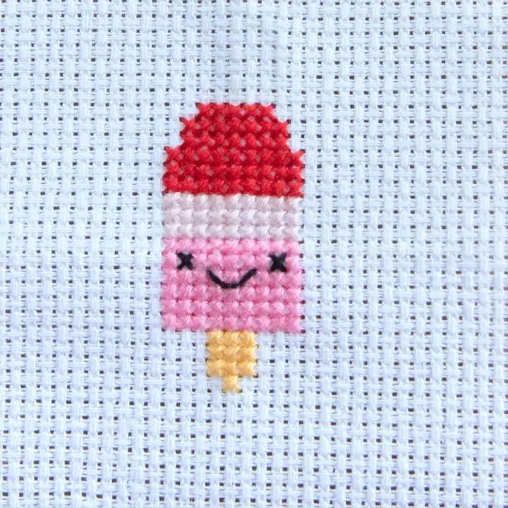 Cross-stitched design of a pink ice cream with a red top on a light blue fabric background.