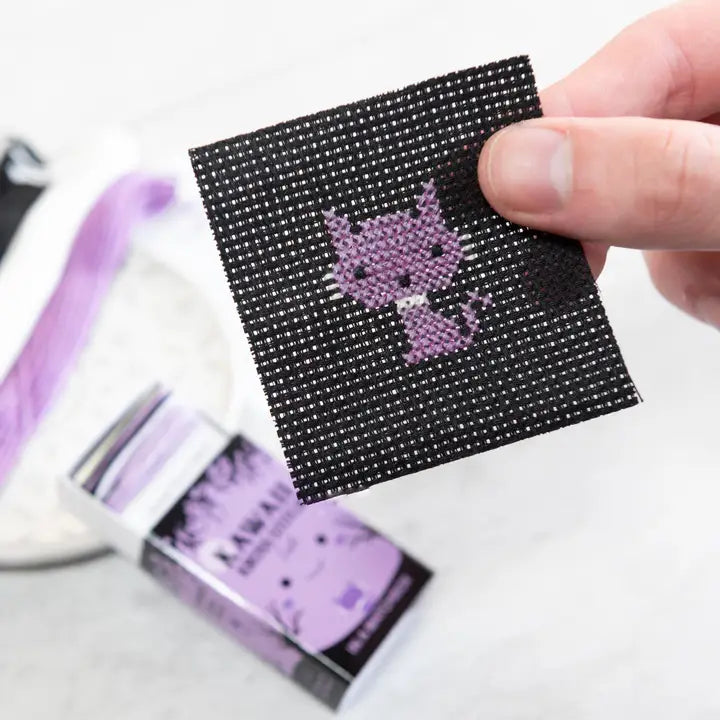 Hand holding a small black square with a purple character design, with a white background.